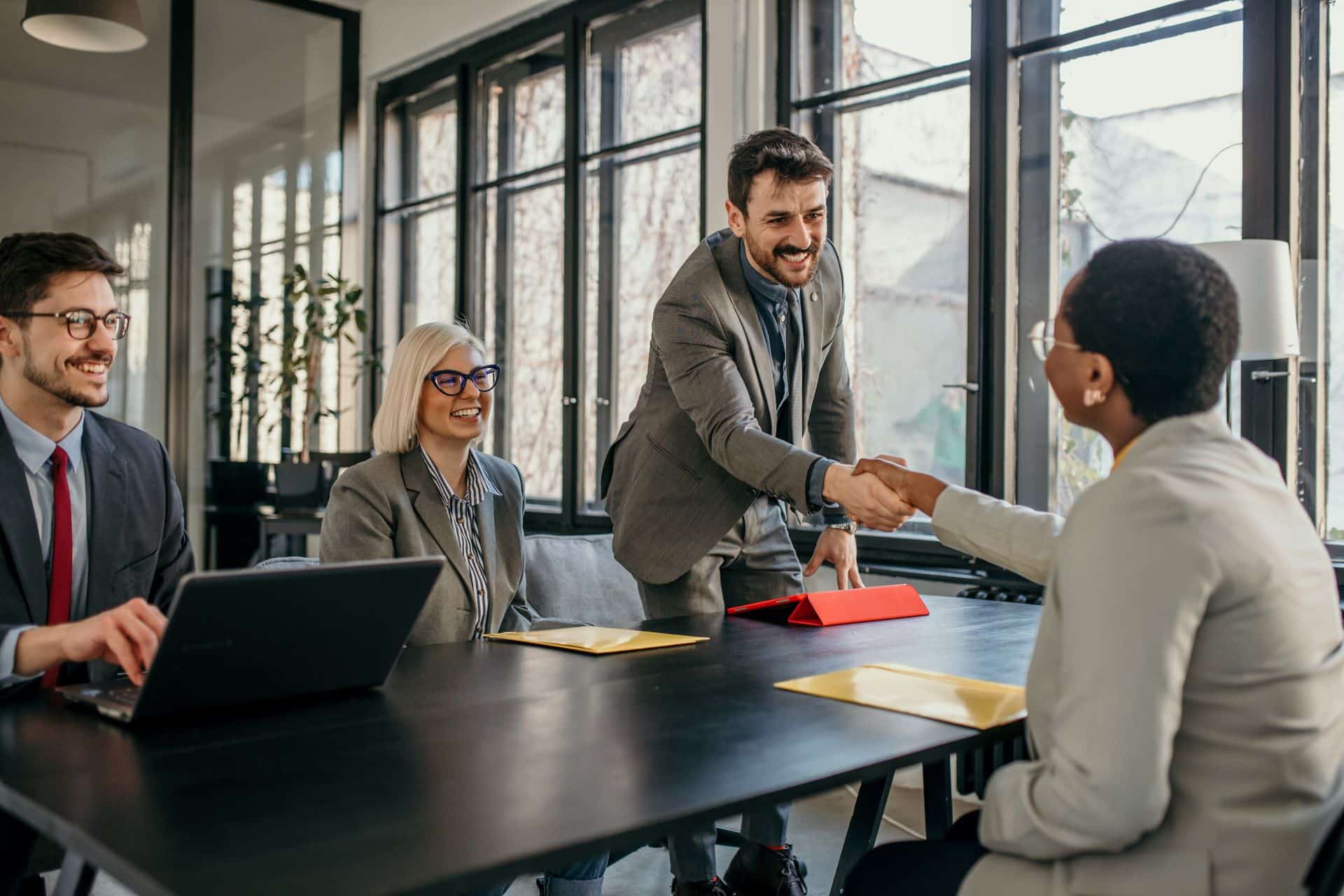 Business-Meeting mit mehreren Teilnehmern, wobei eine Person dem Gegenüber die Hand schüttelt, um eine erfolgreiche Zusammenarbeit zu symbolisieren.