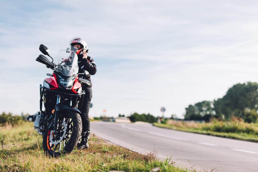 Motorradfahrerin mit rotem Adventure-Bike steht am Straßenrand einer Landstraße bei sonnigem Wetter