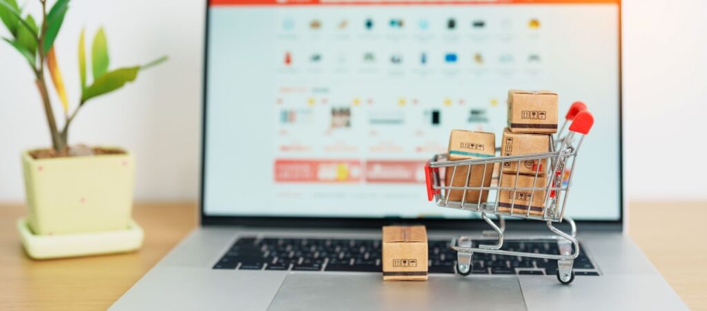 Kleine Kartons im Einkaufswagen auf Laptop vor geoeffneter Verkaufsplattform, Symbol fuer Macbook verkaufen ueber Online-Portale